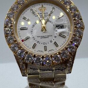 Puede incluir: Un reloj dorado con esfera blanca y bisel engastado con gemas transparentes. La esfera del reloj muestra el texto "ORAN SAPHIRE CRYSTAL TIME UST HIGH QUALITY WATCH AUTOMATIC". La correa tambi&eacute;n es dorada y est&aacute; engastada con gemas m&aacute;s peque&ntilde;as.