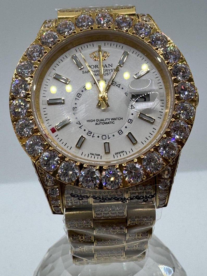 Puede incluir: Un reloj dorado con esfera blanca y bisel engastado con gemas transparentes. La esfera del reloj muestra el texto "ORAN SAPHIRE CRYSTAL TIME UST HIGH QUALITY WATCH AUTOMATIC". La correa tambi&eacute;n es dorada y est&aacute; engastada con gemas m&aacute;s peque&ntilde;as.