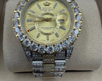 May include: A gold-toned wristwatch with a round face and a diamond-encrusted bezel. The watch face displays the brand name "JORDAN" and the text "SAPPHIRE CRYSTAL TIME JUST". The watch band is silver and gold-toned. The watch is an automatic high-quality watch.