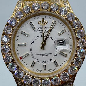 Puede incluir: Reloj de pulsera dorado con esfera blanca, con bisel y correa engastados con diamantes. La esfera del reloj muestra el texto "JORDAN SAPPHIRE CRYSTAL TIME UST" y una visualizaci&oacute;n de la fecha. Es un dise&ntilde;o de reloj autom&aacute;tico de alta calidad.