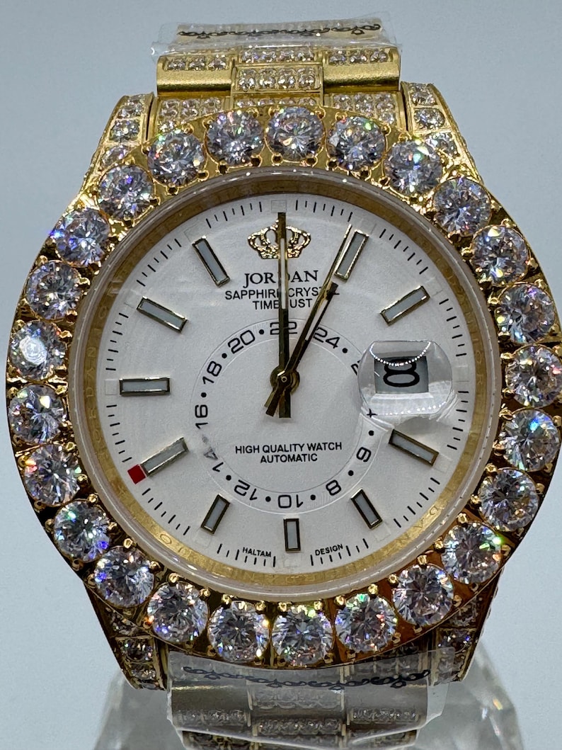 Puede incluir: Reloj de pulsera dorado con esfera blanca, con bisel y correa engastados con diamantes. La esfera del reloj muestra el texto "JORDAN SAPPHIRE CRYSTAL TIME UST" y una visualizaci&oacute;n de la fecha. Es un dise&ntilde;o de reloj autom&aacute;tico de alta calidad.