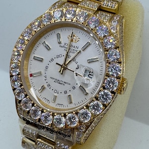 Puede incluir: Un reloj de pulsera dorado con esfera blanca y bisel y correa incrustados con gemas transparentes. La esfera del reloj muestra el texto "JORDAN SAPPHIRE CRYSTAL" y tiene manecillas e indicadores dorados. El reloj es de dise&ntilde;o autom&aacute;tico.