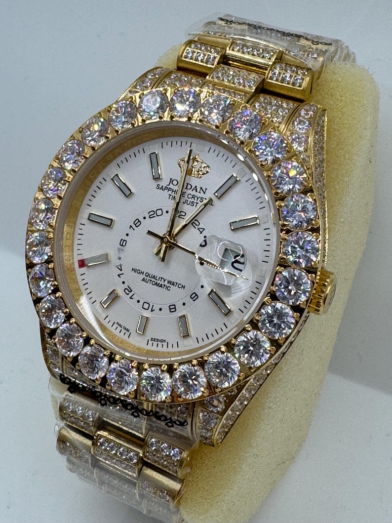 Puede incluir: Un reloj de pulsera dorado con esfera blanca y bisel y correa incrustados con gemas transparentes. La esfera del reloj muestra el texto "JORDAN SAPPHIRE CRYSTAL" y tiene manecillas e indicadores dorados. El reloj es de dise&ntilde;o autom&aacute;tico.
