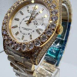 Puede incluir: Un reloj de pulsera dorado con esfera blanca, con bisel y brazalete engastados con diamantes. La esfera muestra la hora y el texto "JORDAN SAPPHIRE CRYS TIM JUST HIGH QUALITY WATCH AUTOMATIC". El reloj se exhibe sobre un soporte transparente.