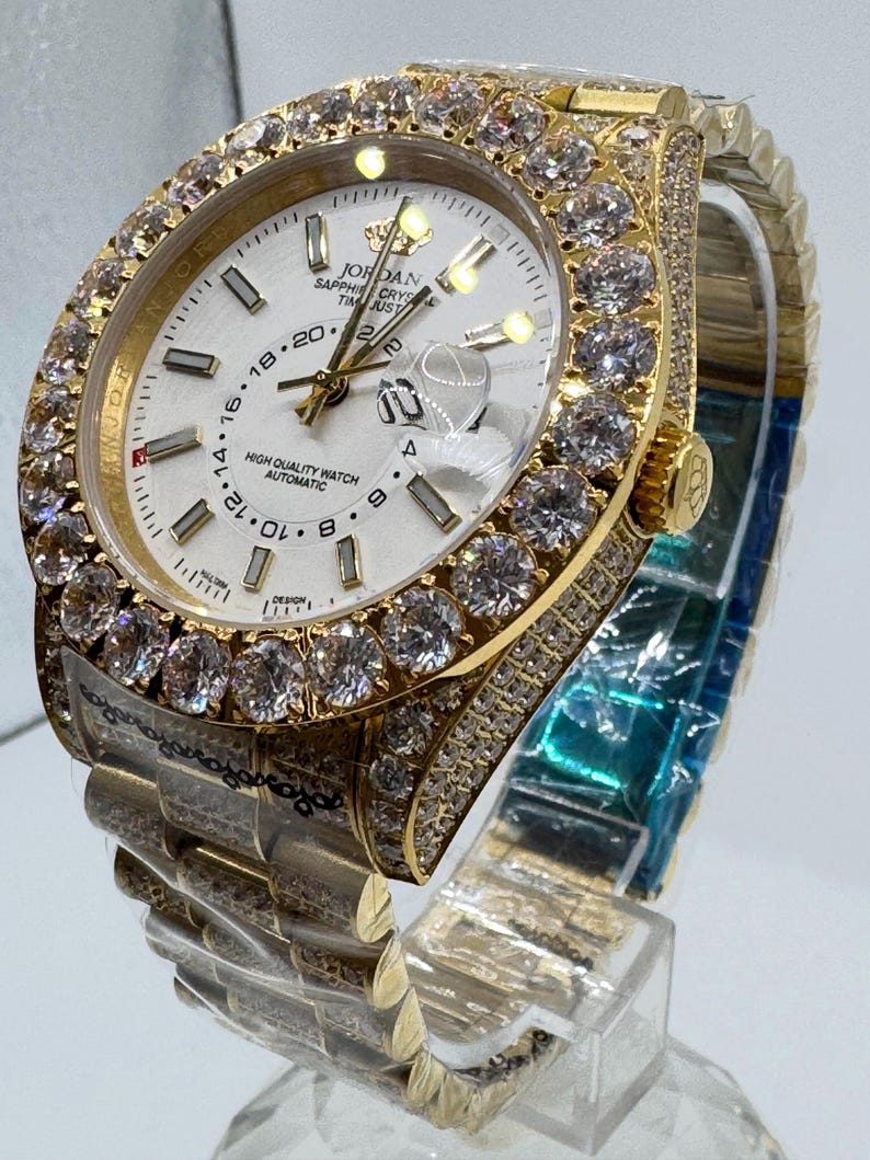 Puede incluir: Un reloj de pulsera dorado con esfera blanca, con bisel y brazalete engastados con diamantes. La esfera muestra la hora y el texto "JORDAN SAPPHIRE CRYS TIM JUST HIGH QUALITY WATCH AUTOMATIC". El reloj se exhibe sobre un soporte transparente.