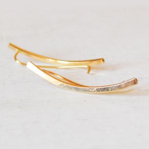 May include: A pair of gold ear cuffs with a curved, textured bar design. The cuffs are made of a smooth, shiny metal.