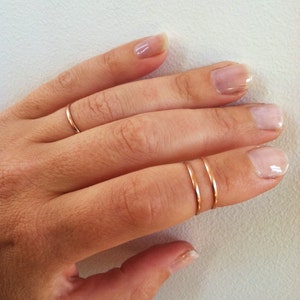 May include: A hand with three thin, rose gold rings. Two rings are stacked on one finger, and a single ring is on another. The rings are simple and minimalist in design.