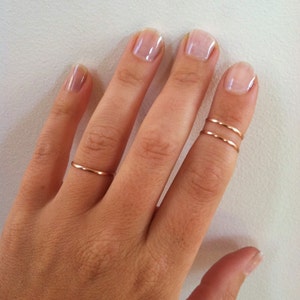 May include: A hand wearing delicate, thin, gold rings. One ring is a single band, while another finger features two stacked bands. The rings have a simple, minimalist design. The background is a light, neutral color.