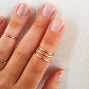 May include: A hand wearing multiple thin gold rings on the middle finger. The nails are painted a light pink color.