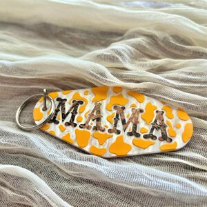 May include: Oval keychain with the word "MAMA" in brown camouflage letters. The keychain has a white background with yellow cow spots and a silver key ring.