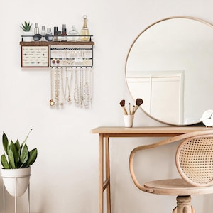 May include: A wall-mounted jewelry organizer with a dark wood shelf and black metal frame. The organizer displays necklaces, earrings, and other accessories. Various beauty products and a small plant sit on the shelf above. A round mirror and vanity are in the background.