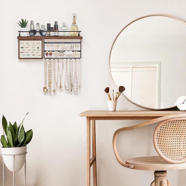 May include: A wall-mounted jewelry organizer with a dark wood shelf and black metal frame. The organizer displays necklaces, earrings, and other accessories. Various beauty products and a small plant sit on the shelf above. A round mirror and vanity are in the background.