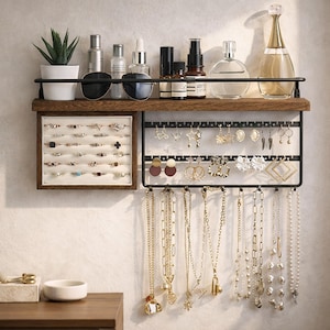 May include: A wall-mounted jewelry organizer with a dark wood shelf and black metal frame. The shelf holds a small potted plant, sunglasses, and various beauty products. The organizer displays rings, earrings, and necklaces.