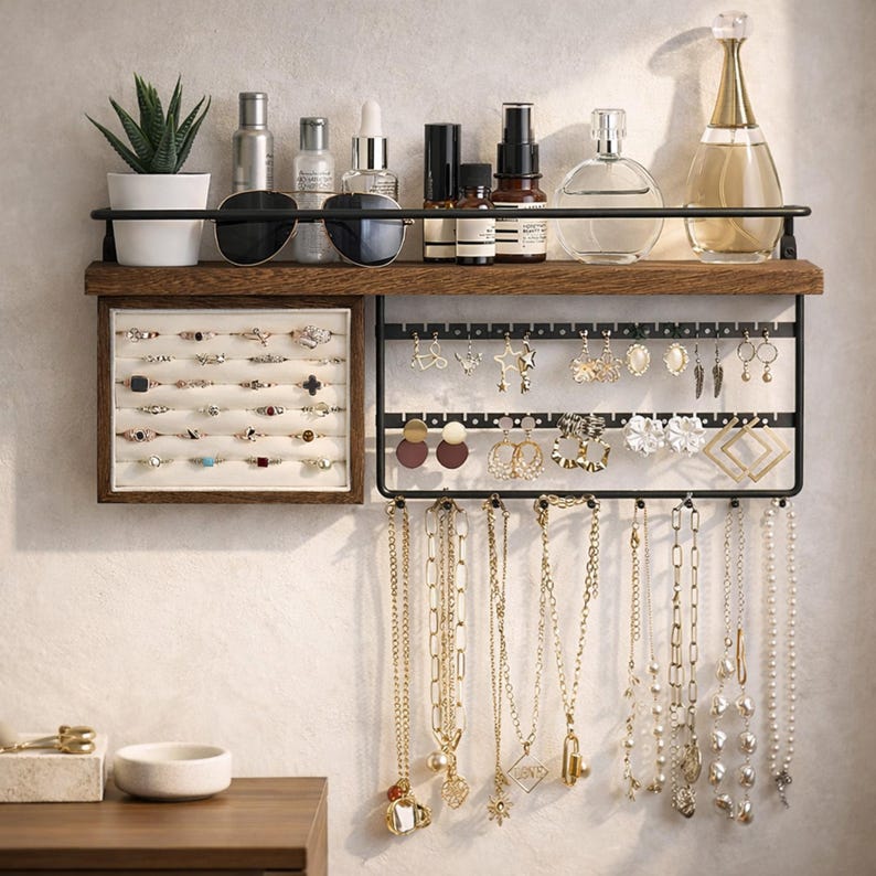May include: A wall-mounted jewelry organizer with a dark wood shelf and black metal frame. The shelf holds a small potted plant, sunglasses, and various beauty products. The organizer displays rings, earrings, and necklaces.