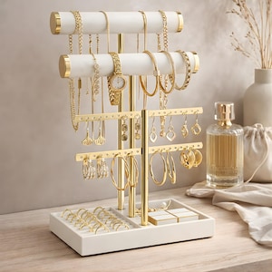 May include: A white and gold jewelry organizer displaying various gold necklaces, bracelets, earrings, and a watch. The organizer has multiple tiers and a base with ring holders and small compartments. The jewelry is arranged on the organizer.