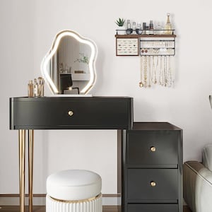 May include: A black vanity with gold legs, a white stool, and a large, illuminated, abstract-shaped mirror. A wall-mounted jewelry organizer displays necklaces and earrings. Makeup brushes and bottles are on the vanity.