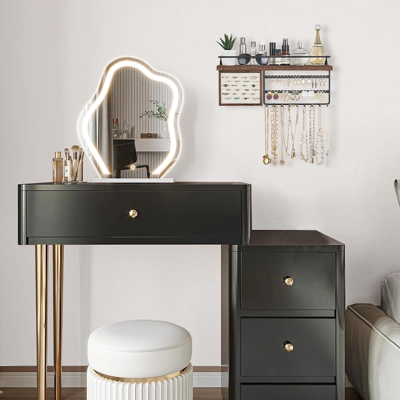 May include: A black vanity with gold legs, a white stool, and a large, illuminated, abstract-shaped mirror. A wall-mounted jewelry organizer displays necklaces and earrings. Makeup brushes and bottles are on the vanity.