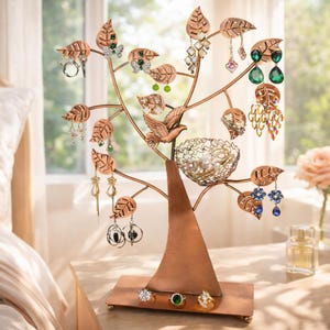May include: A copper-toned metal jewelry organizer shaped like a tree with leaf-shaped branches for hanging earrings. The tree features a bird figure, a wire nest for small items, and a rectangular base holding three rings with gemstones.