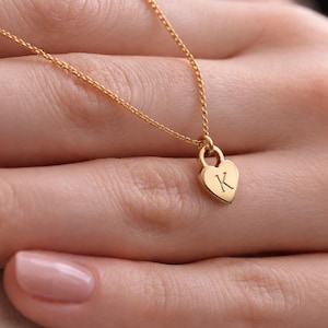 Puede incluir: Un collar dorado con una cadena fina y un colgante en forma de corazón. El colgante tiene grabada la letra "K". El collar se muestra en la mano de una persona.