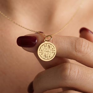 May include: A gold-colored pendant necklace featuring a circular charm with an intricate, woven design. The pendant hangs from a delicate chain. The necklace is held by a hand with dark red nail polish.