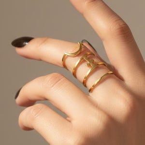 May include: A gold-colored finger ring with multiple connected bands. The ring is worn on the middle finger and extends to the knuckle. The ring has a unique, modern design. The background is a solid, neutral color.