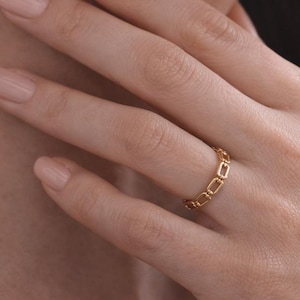 May include: A gold ring with a repeating rectangular design is worn on a person's finger. The ring is the focal point, with the person's hand and arm in the foreground. The background is blurred, with a dark color.