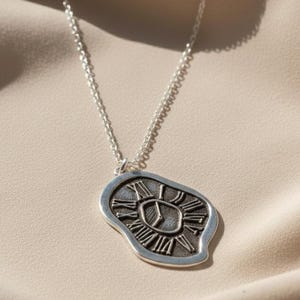 May include: A silver-toned necklace with a pendant shaped like a melting clock. The pendant features Roman numerals and a clock face design. The necklace chain is delicate and hangs on a beige fabric background.