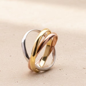 May include: A set of three interlocking rings in silver, gold, and rose gold. The rings are intertwined, creating a unique and elegant design. The jewelry is displayed on a neutral surface.