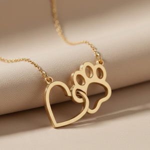 May include: A gold-colored necklace featuring a heart and a paw print pendant. The pendant is connected to a delicate chain. The necklace is displayed against a neutral background, highlighting the jewelry's design and color.