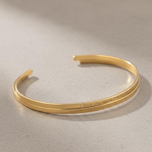 May include: A gold-coloured cuff bracelet with the inscription "I love you mom" and a date. The bracelet has a simple, elegant design and is made of a shiny metal. The bracelet is open-ended.