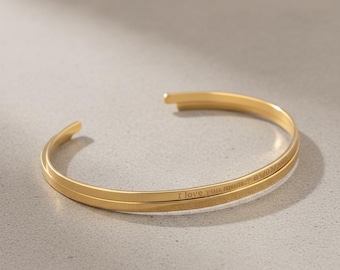 May include: A gold-colored cuff bracelet with the inscription "I love you mom" and a date. The bracelet has a simple, elegant design and is made of a shiny metal. The bracelet is open-ended.