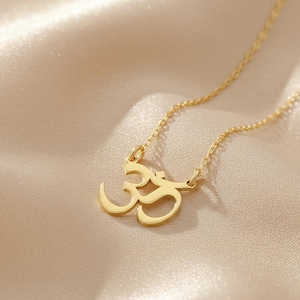 May include: A gold-colored necklace featuring the Om symbol pendant. The pendant is a stylized script, suspended from a delicate chain. The necklace is displayed on a soft, textured, beige fabric.
