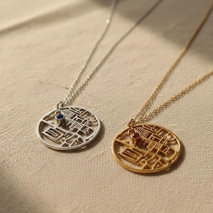 May include: Two necklaces with circular pendants featuring a city map design. One necklace is silver with a blue gemstone, the other is gold with a red gemstone. Both are on delicate chains, set against a neutral background.