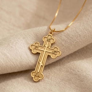May include: A gold-colored Orthodox cross pendant with a delicate chain. The cross features intricate detailing and religious inscriptions. The pendant is displayed on a beige fabric, highlighting its design and craftsmanship. A piece of religious jewelry.