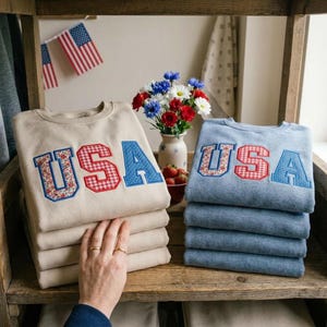 May include: Stacks of beige and blue sweatshirts with the word "USA" in red gingham, floral, and blue polka dot letters. American flags and a vase of flowers are in the background. The sweatshirts are neatly folded on a wooden shelf.