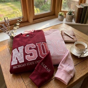 May include: Two sweatshirts, one maroon and one pink, with embroidered lettering. The maroon sweatshirt says "NSU MICHIGAN STATE UNIV" with a lace and gingham design. The pink sweatshirt says "NSU UNIVERSITY". Two matching maroon and pink cuffs are also present.