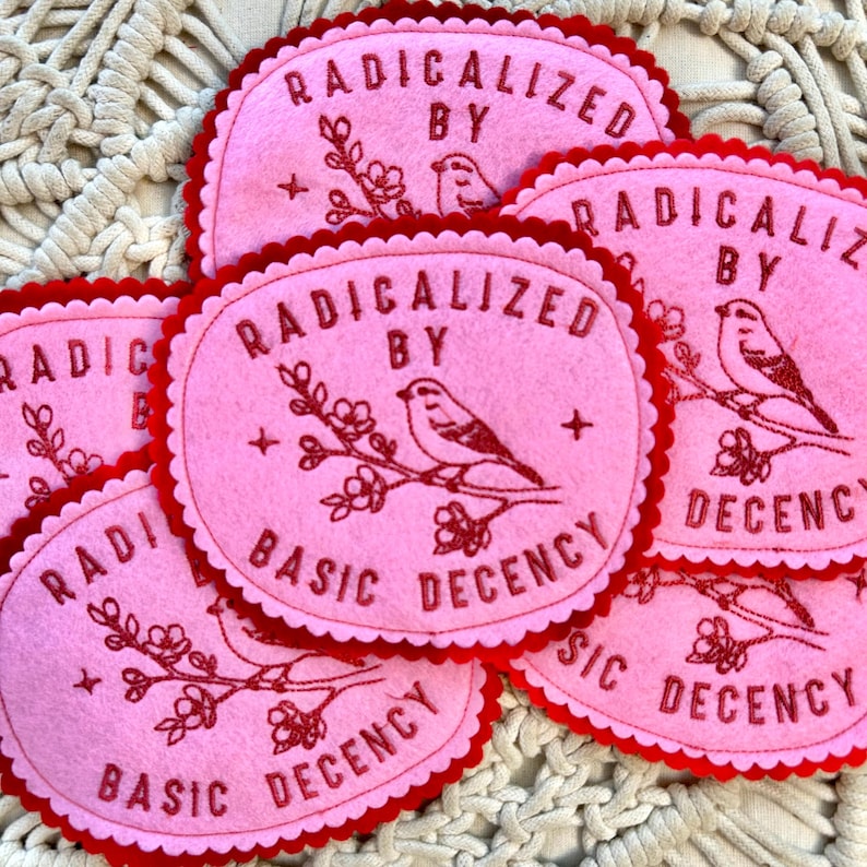 K&ouml;nnte beinhalten: Rosa Filzuntersetzer mit rotem, gewelltem Rand. Jeder Untersetzer tr&auml;gt den Text "RADICALIZED BY BASIC DECENCY" und einen roten, gestickten Vogel auf einem bl&uuml;henden Zweig. Ein Set dekorativer Getr&auml;nkeuntersetzer.