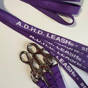 May include: Purple dog leash with the text "A.D.H.D. LEASH" in white. The leash has silver-colored metal clasps. The leash is made of a woven material.