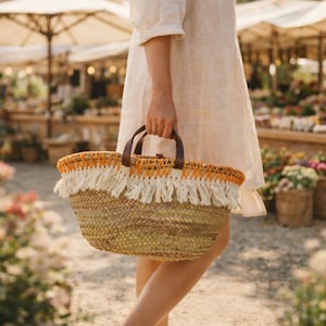 Pode incluir: Uma cesta de palha tecida com uma alça de couro marrom e acabamento de franjas laranja e branco. A cesta está sendo carregada ao ar livre, com um fundo desfocado de flores e guarda-sóis de mercado.
