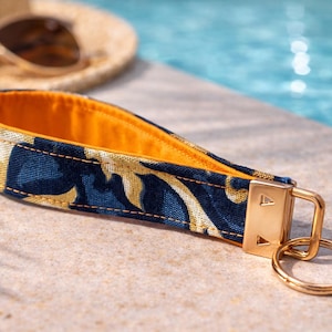 Pode incluir: Um porta-chaves com um padrão azul marinho e dourado, com um interior laranja brilhante. O porta-chaves tem um fecho e um porta-chaves de metal dourado. O fundo inclui óculos de sol e uma piscina.