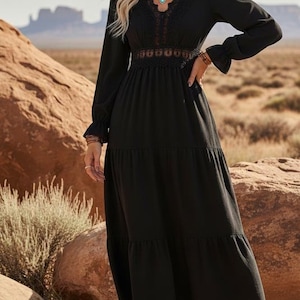 May include: A black maxi dress with long sleeves, a tiered skirt, and lace detailing. The outfit includes a tan hat and brown cowboy boots. The backdrop is a desert scene with rock formations and a clear sky.