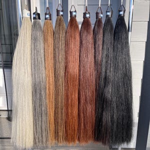 Könnte beinhalten: Eine Präsentation von acht Haarverlängerungen in verschiedenen Farbtönen, von hellblond bis schwarz. Jede Verlängerung ist an einer kleinen, dunkel gefärbten Basis befestigt und hängt vertikal an einem Metallgestell. Die Farben umfassen Grau-, Braun- und Rotbrauntöne.