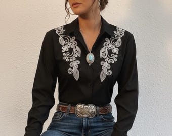 May include: A black long-sleeve shirt with white floral embroidery, paired with blue jeans and a brown belt with a silver buckle. A beige cowboy hat and a turquoise pendant necklace complete the Western-inspired outfit.