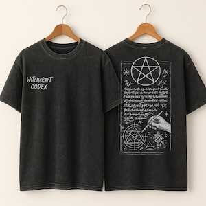 Puede incluir: Camisetas de color gris oscuro con el texto "Witchcraft Codex" en la parte delantera. La parte trasera de la camiseta presenta un pentagrama, texto manuscrito e ilustraciones de una mano dibujando un círculo con un bolígrafo. Las camisetas están colgadas en perchas de madera.