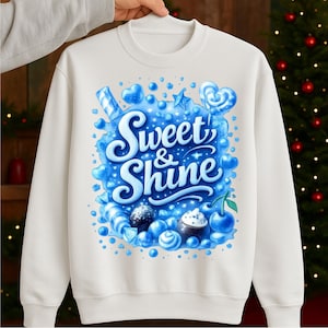 May include: A white sweatshirt featuring a blue graphic with the words "Sweet & Shine". The design includes blue candies, hearts, and other sweets. The sweatshirt is held up by a person.