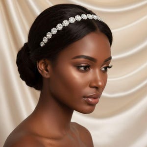 May include: A sparkling silver headband with round, clear crystals. The headband is worn on a person with dark hair styled in a low bun. The background is a soft, cream-colored fabric.