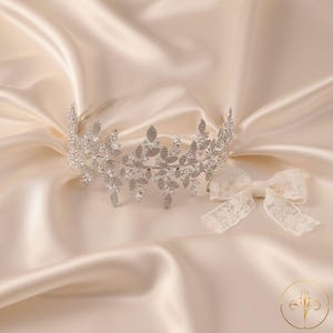 Puede incluir: Una corona nupcial plateada con dise&ntilde;o de hojas, adornada con cristales brillantes. Un lazo de encaje marfil se coloca junto a la corona sobre un fondo satinado. La corona es un hermoso accesorio para bodas y ocasiones especiales.