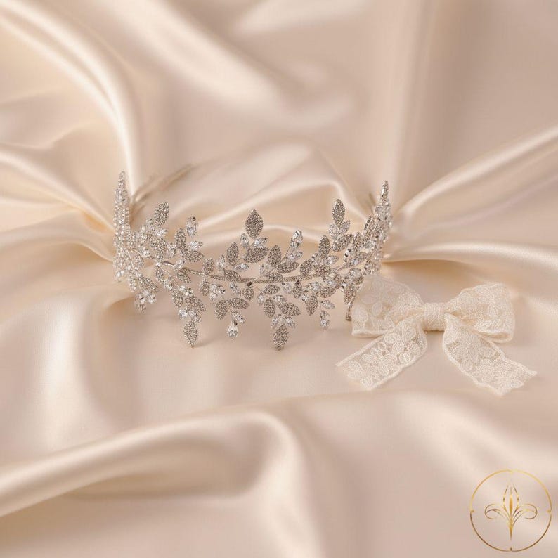 Puede incluir: Una corona nupcial plateada con dise&ntilde;o de hojas, adornada con cristales brillantes. Un lazo de encaje marfil se coloca junto a la corona sobre un fondo satinado. La corona es un hermoso accesorio para bodas y ocasiones especiales.