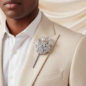 May include: A silver and crystal floral lapel pin with a pearl centre, attached to a light grey fabric base. The pin is affixed to a cream-coloured suit jacket, paired with a white dress shirt.