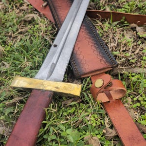 May include: A medieval-style sword with a silver blade and a brass crossguard. The handle is wrapped in dark red leather. The sword rests on a brown leather sheath with a matching belt, all set against a backdrop of green grass.